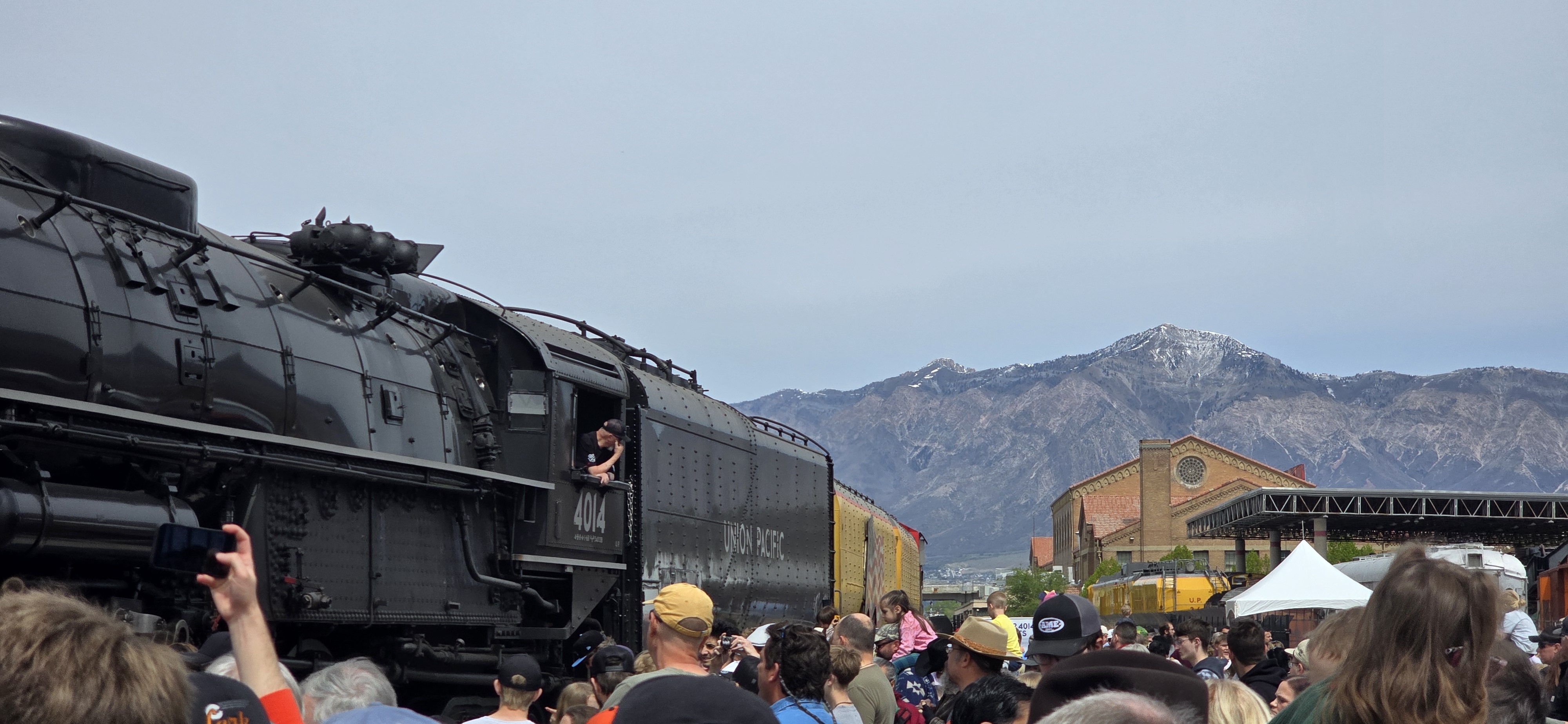 A crowd gathered around a historic Union Pacific steam locomotive, many holding up phones to take photos, with mountains in the background.